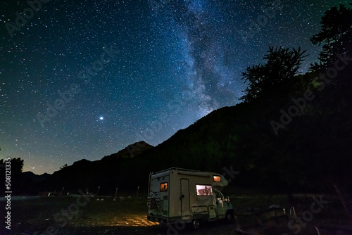 Camper van under panoramic night sky in the Alps. The Milky Way galaxy arc and stars over illuminated motorhome. Camping freedom in unique landscape.