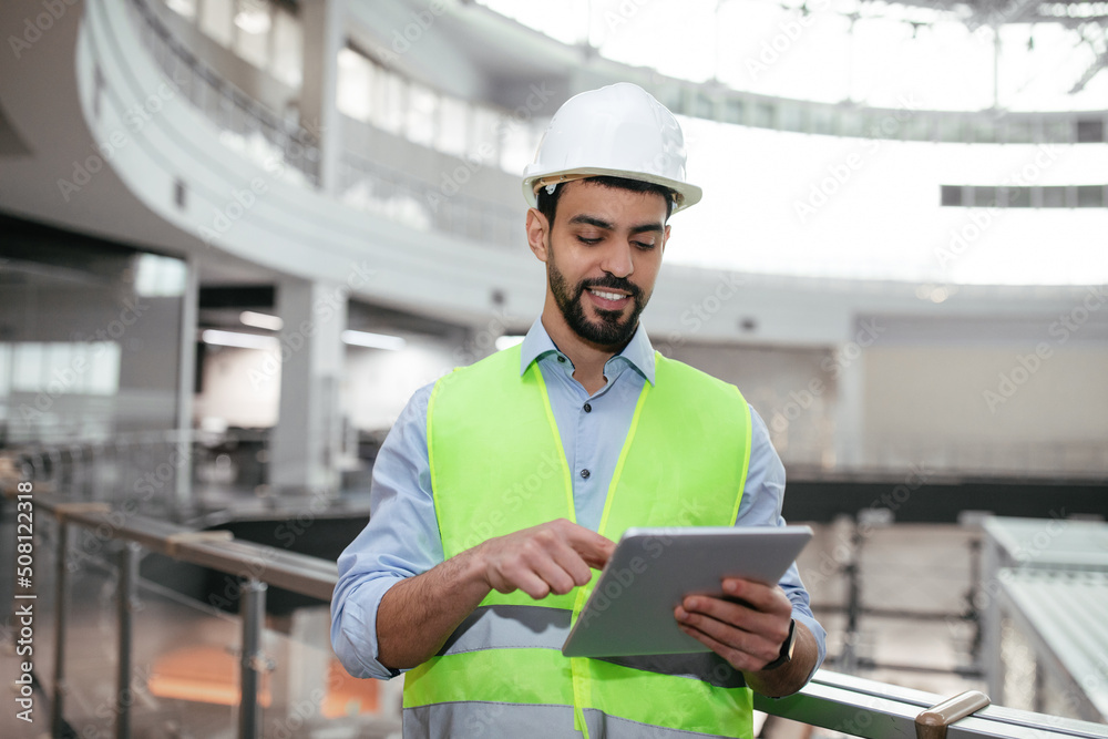 Cheerful young muslim engineer builder with beard in protective clothing typing on tablet, checking work Cheerful young muslim engineer builder with beard in protective clothing typing on tablet, checking work