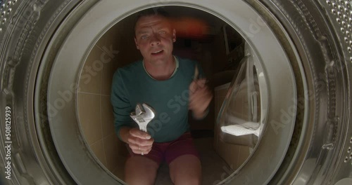 Cheerful man in a washing machine, holding a key and pliers with his hands. He intends to make a belt device, dilettante. The head looks from the inside. Joy in doing work. photo