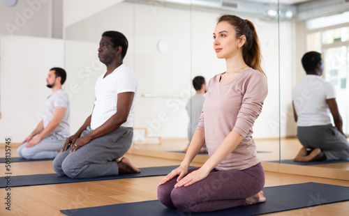 Relaxed young woman practicing Vajrasana asana during group training in fitness studio, sitting on her heels on mat with hands resting on knees