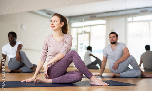 Positive young woman doing yoga with group in fitness studio, sitting in twisting asana Matsyendrasana ..