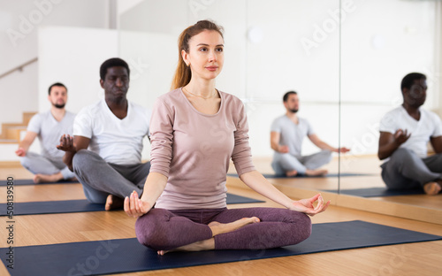 Sporty slim young woman sitting in Padmasana yoga position with her hands resting on knees in mudra during group training in gym..