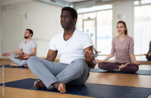 Focused adult african american man meditating in modern yoga studio, sitting in lotus position with hands on knees with fingers folded in mudra