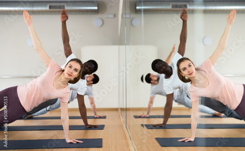 Concentrated young woman practicing power yoga during group training in fitness studio, standing in side plank pose Vasisthasana near mirror with reflection