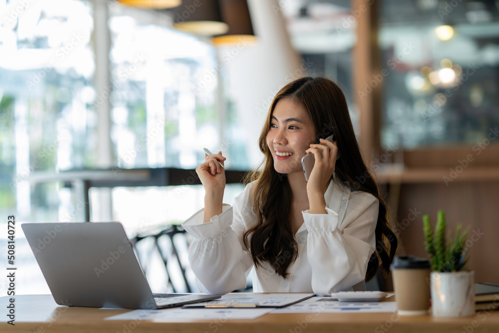 Happy business asian manageress working at her desk in the office taking a call on her mobile phone while writing notes on a notepad Happy business asian manageress working at her desk in the office taking a call on her mobile phone while writing notes on a notepad