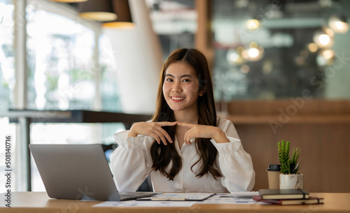 Happy Asian businesswoman laughing sitting at the work desk with laptop, a cheerful smiling female employee having fun feeling joy and positive emotion express sincere laughter at office workplace