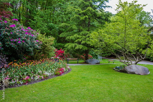 Resting place at Victoria Hill Park of New Westminster City. Bench is located under the canopy of spreading tree on a green lawn with flower beds among flowering shrubs,   British Columbia, Canada