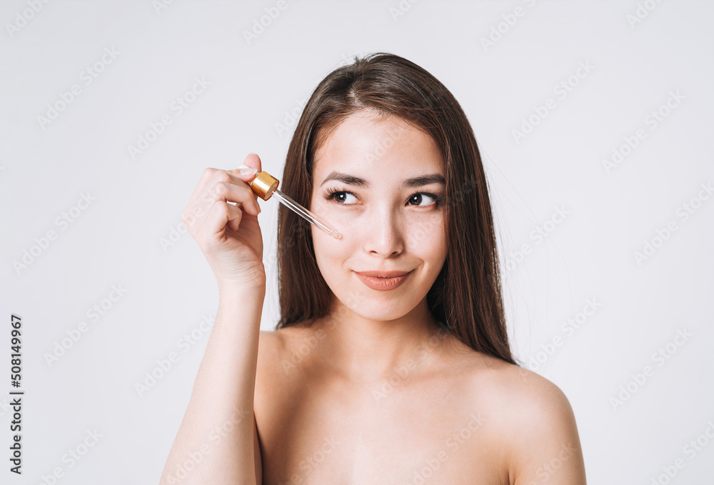 Beauty portrait of happy smiling asian woman with dark long hair with pipette with organic oil on clean fresh skin face and hands on white background isolated