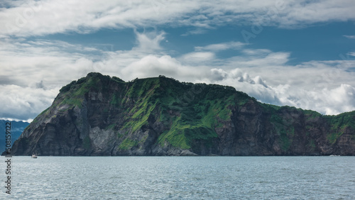 The picturesque coast of Kamchatka against the background of blue sky and clouds. Green vegetation is visible on the steep slopes of the rocks. The boat sails on the Pacific Ocean. Avacha Bay