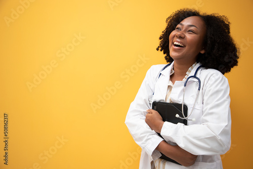 Charming of smiling doctor posing on a yellow background holding her tablet in her hands and a stethoscope around her neck. Health care and technology concept