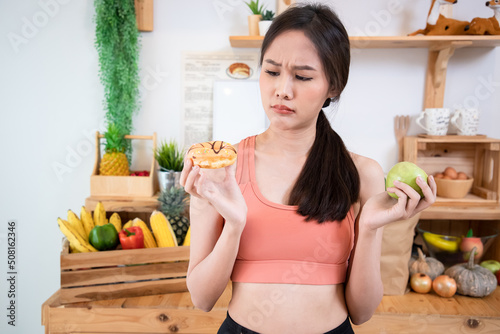 Sport girl dieting or good healthty concept. Young woman rejecting Junk food or unhealthy food such as donut or dessert and choosing healthy food such as fresh fruit or vegetable in kitchen. photo