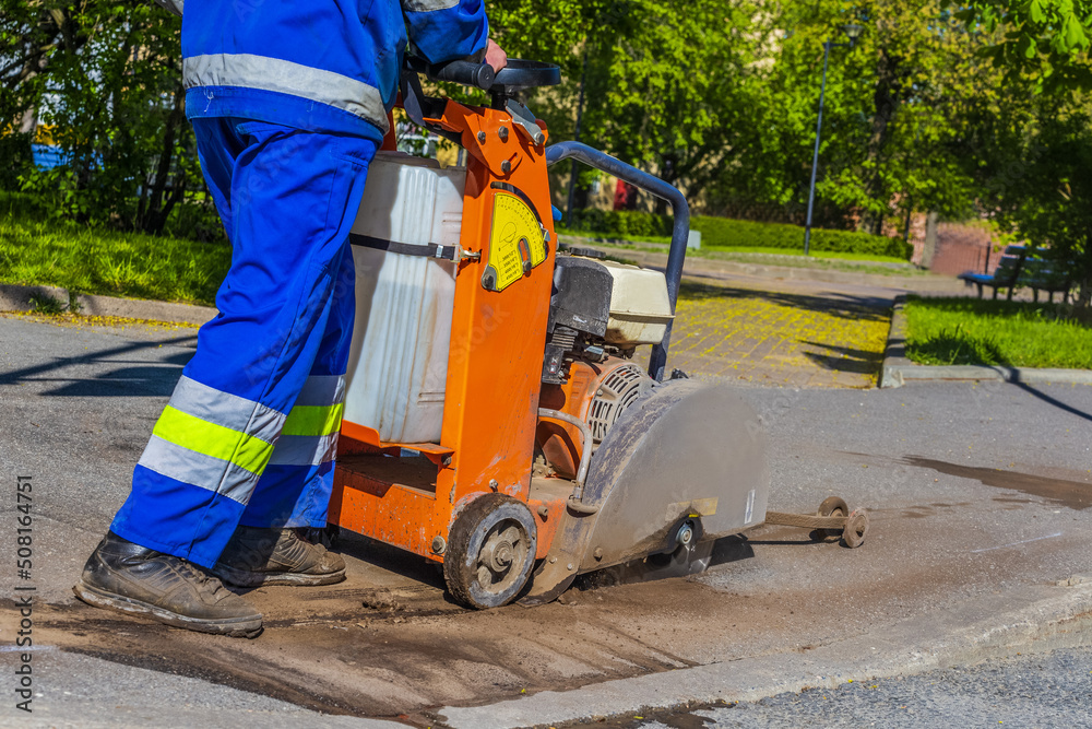 A road worker in overalls uses a portable asphalt cutter to cut asphalt with a diamond blade to repair part of the roadway. Cutting road works with a gasoline-powered angle grinder. Road repairs.