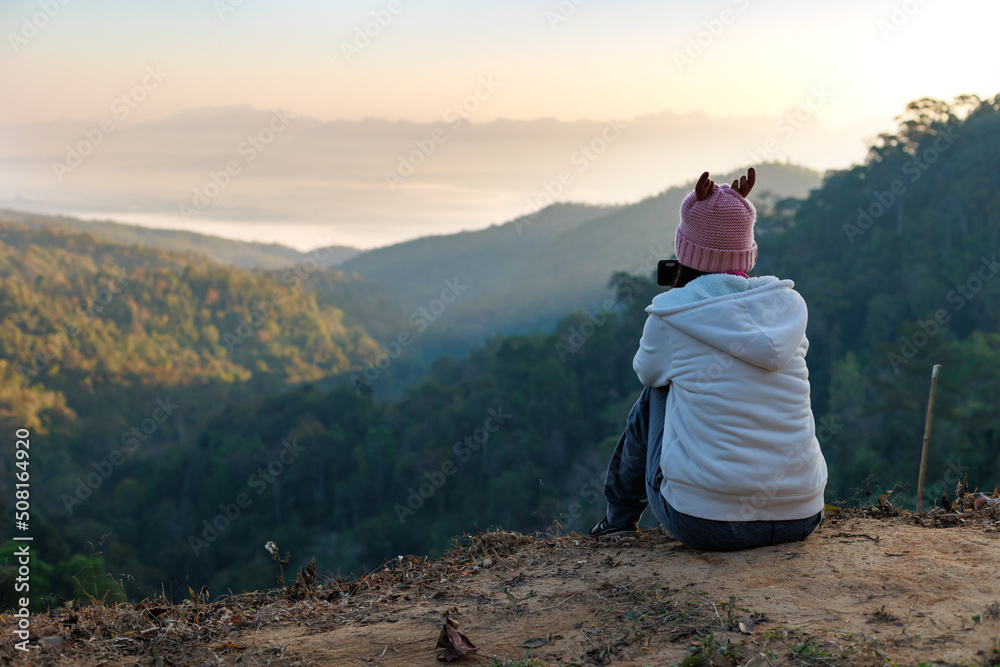 Rear view of asian woman sitting on the ground looking on the view of beautiful nature on the top of mountain. People enjoying with panoramic view in the morning time.