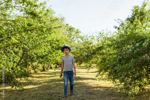 A cheerful kid in jeans, a T-shirt and a blue hat is playing in the garden among the apple trees.