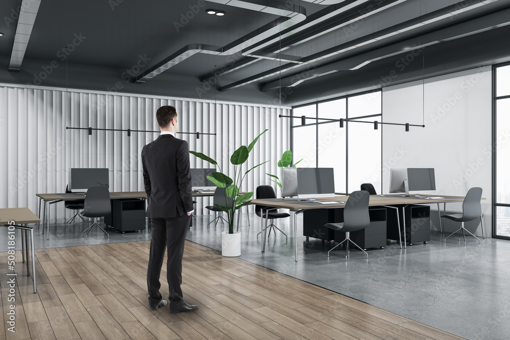 Attractive thoughtful young european man standing in wooden and concrete office interior with furnituire and window with city view. CEO, executive and consulting concept.