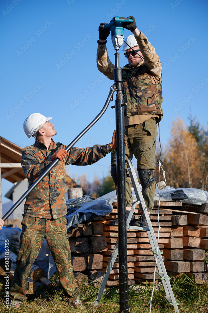 Male workers building pile foundation for wooden frame house. Men builders in white safety helmets drilling piles into the ground on blue sky background.