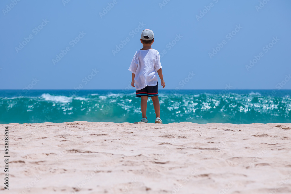 A little boy looks into the blue endless ocean on a tropical beach