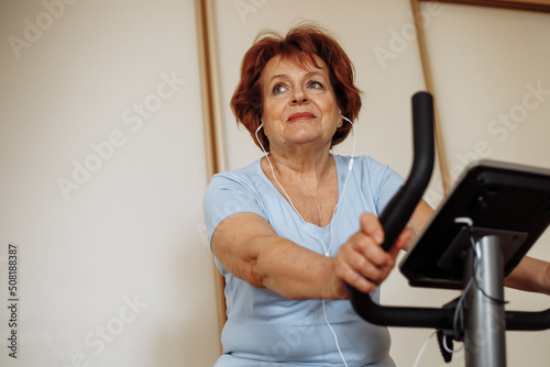Portrait of elderly woman with dark hair  make-up wearing blue T-shirt  sitting on stationary bicycle  doing exercises.