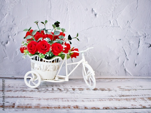 Artificial red rose flowers in bike toy on table bouquet bucket Bicycle with soft tone festive texture background cement wallpaper copy space lettering Valentine's day romantic love birthday card 