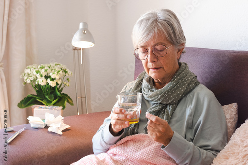 Elderly adult caucasian woman with cold and fever symptoms such as seasonal flu or pollen allergy while taking a medicine with a glass of water