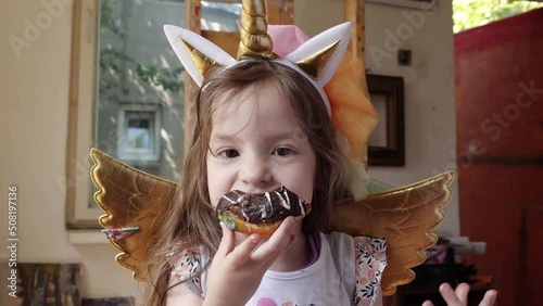 A cute little girl at a party in a unicorn costume bites off a sweet donut with chocolate cream