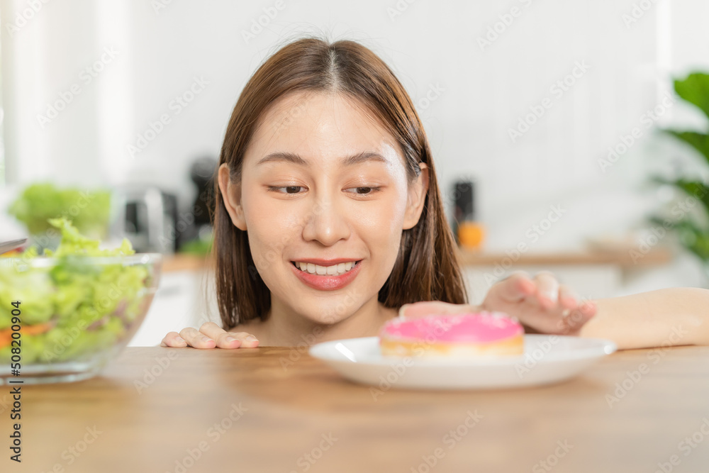 Diet, Dieting asian young woman or girl smile, confused choose, choice eating green salad or pink sweet doughnut at home, eat food for good healthy, health when hungry. Weight loss person concept. Diet, Dieting asian young woman or girl smile, confused choose, choice eating green salad or pink sweet doughnut at home, eat food for good healthy, health when hungry. Weight loss person concept.
