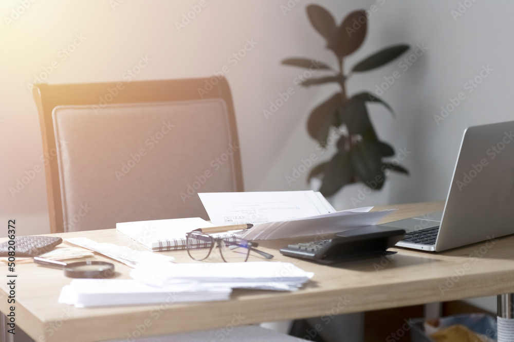 Fototapeta premium Office of an accountant or businessman. On the desktop are papers, a calculator, a laptop, glasses and a notepad. Sunlight shines through the window onto a desktop with papers and ficus in the backgro