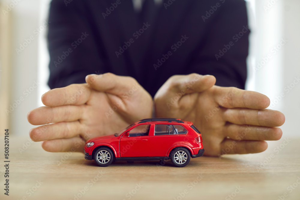 Human hands protecting small red toy car. Close up of little automobile model on table and hands of businessman protecting it. Car insurance, auto safety, warranty, protection concepts