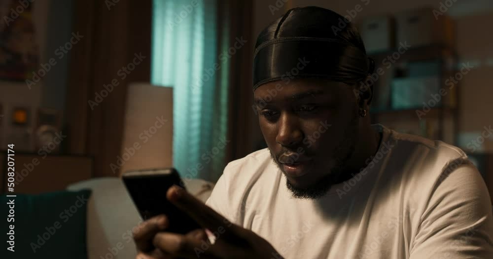 Young African-American man holding smartphone, texting or playing mobile games, sitting on couch at home, uses social media app to send messages browses websites on phone. Young African-American man holding smartphone, texting or playing mobile games, sitting on couch at home, uses social media app to send messages browses websites on phone.
