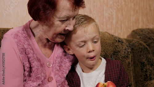 A close-up of a grandmother and grandson sitting together on the couch, a boy eating an apple that his grandmother gave him.