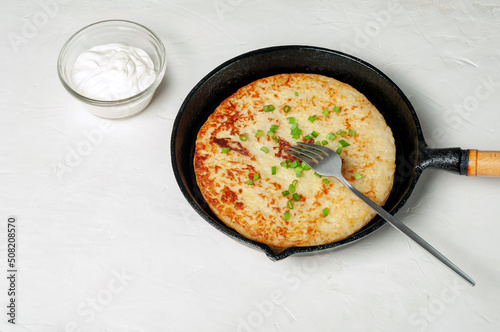 Swiss traditional dish is a potato pancake in a frying pan with a fork, decorated with green onions. there is a gravy boat with white cream sauce nearby.