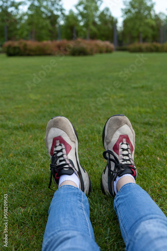 photo of women's legs sitting in jeans and stylish leather sneakers on the grass, in the park, on a sunny day. Street photography in the first person © Lina Solntseva 