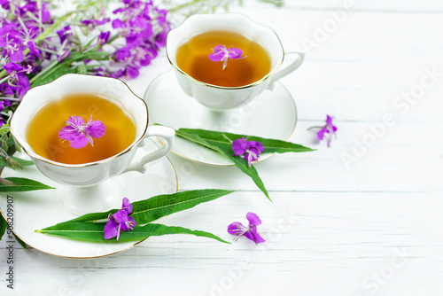 Two cups of infusion (drink, decoction) of ivan-tea on a white wooden background. Grass and flowers of the plant kiprey, ivan-grass, epilobium. Space for the text.