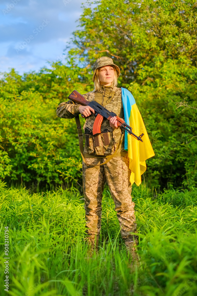 Full length Armed with assault rifle determined female soldier in camouflage uniform and covered with Ukrainian flag shoulder looking away. Ukrainian woman patriot. Vertical orientation