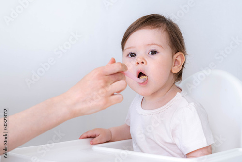 Baby girl eating blend mashed food sitting, on high chair, mother feeding child, hand with spoon for vegetable lunch, baby weaning, first solid food for young kid.