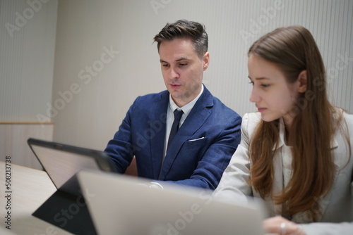 Image of young businessmen and colleague discussing document in laptop and touchpad at meeting