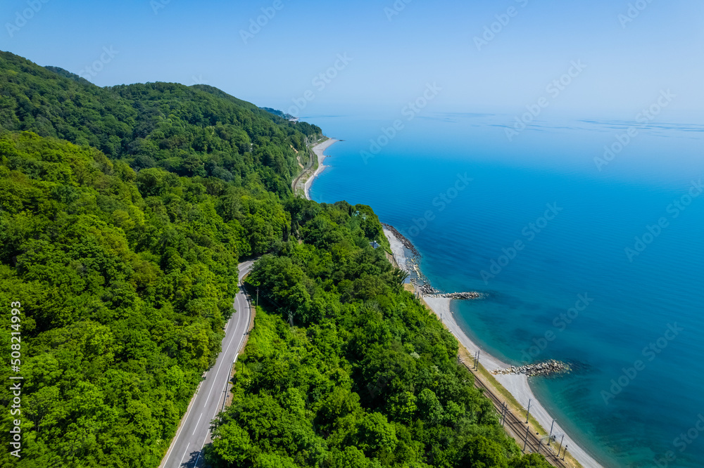 Asphalt road is meandering between blue sea and green mountains. Aerial view of car driving along the winding mountain road in Sochi, Russia.