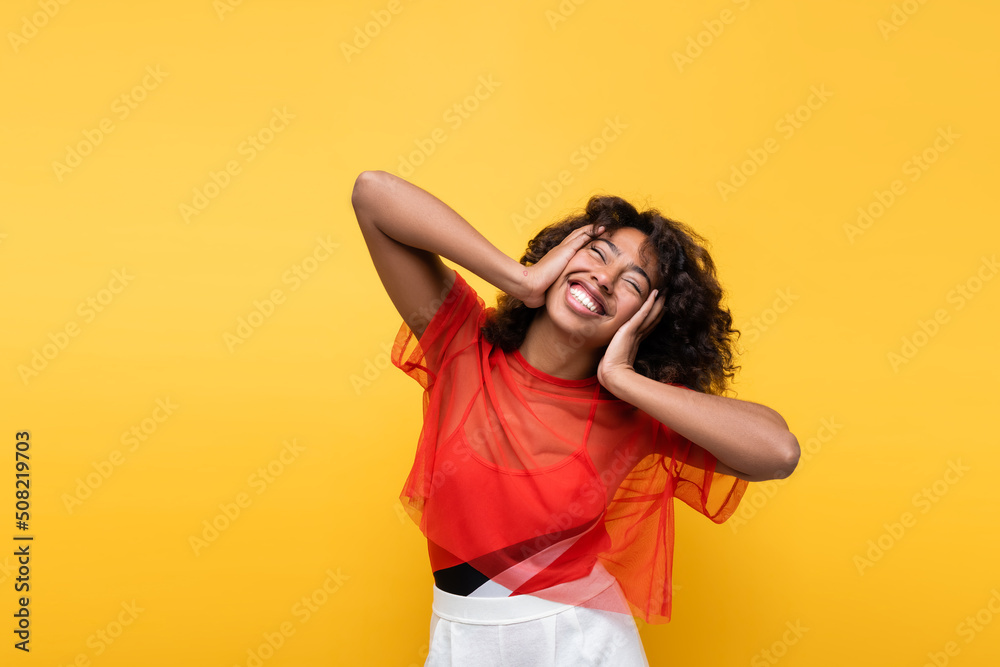 happy african american woman touching face while smiling with closed eyes isolated on yellow. happy african american woman touching face while smiling with closed eyes isolated on yellow.