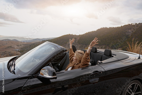 Fotografie, Obraz Young happy smiling female traveling in convertible car by mountainside holding