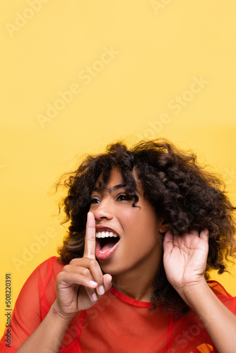 excited african american woman holding hand near ear and showing hush sign isolated on yellow.
