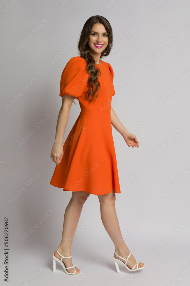 Young woman in orange cocktail dress and high heels sandals is walking and smiling. Young woman in orange cocktail dress and high heels sandals is walking and smiling.