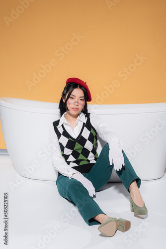Fashionable asian woman in beret and gloves sitting near bathtub on orange background.
