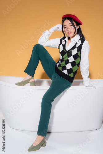 Stylish asian model in beret and gloves posing on bathtub on orange background.