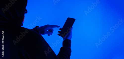 Anonymous hacker and face mask with smartphone in hand. Man in black hood shirt trying to hack personal data from mobile phone. Represent cyber crime data hacking or personal data stealing concept.