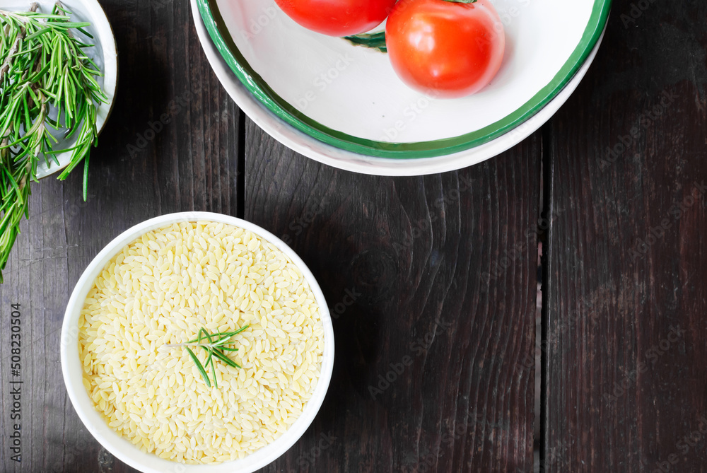 Raw risoni pasta in a white bowl with rosemary sprig and ingredients on a wooden table. The concept of Italian cuisine. Horizontal orientation. Top view. copy space. Raw risoni pasta in a white bowl with rosemary sprig and ingredients on a wooden table. The concept of Italian cuisine. Horizontal orientation. Top view. copy space.