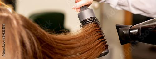 A hairdresser is drying long brown hair with a hairdryer and round brush in a beauty salon