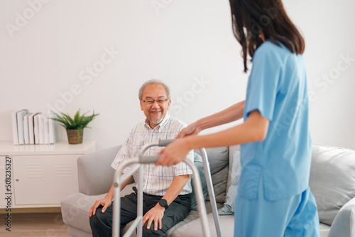 Young asian female caregiver helping older male to walk with walker at home.