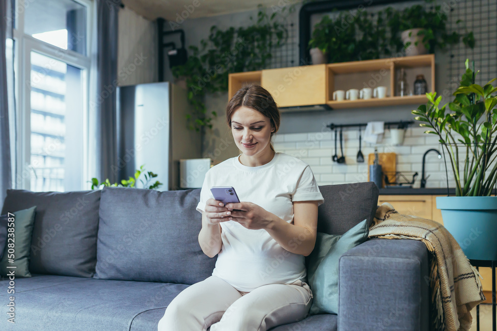 Maternity leave. Pregnant beautiful woman sitting on the couch at home, holding a mobile phone, corresponding in parent chats, typing messages, calling, watching, receiving good news. Smiling, happy Maternity leave. Pregnant beautiful woman sitting on the couch at home, holding a mobile phone, corresponding in parent chats, typing messages, calling, watching, receiving good news. Smiling, happy