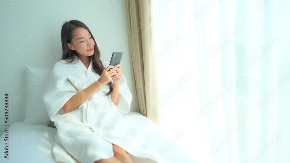 Young Beautiful Female in Bathrobe Texting on Mobile Phone by Window on Bright Morning Young Beautiful Female in Bathrobe Texting on Mobile Phone by Window on Bright Morning