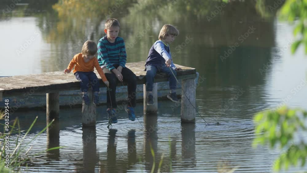 Children sit on wooden pontoon and play with stick on water lake, fishing rod Children sit on wooden pontoon and play with stick on water lake, fishing rod